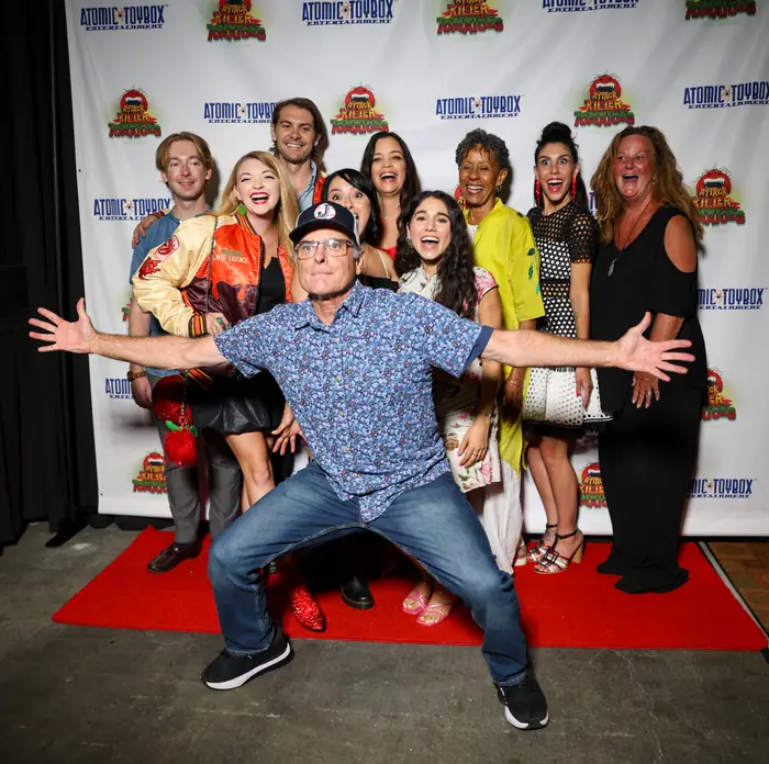 Cast and crew crowded together on the red carpet, one guest crouching front and centre with arms flung wide.