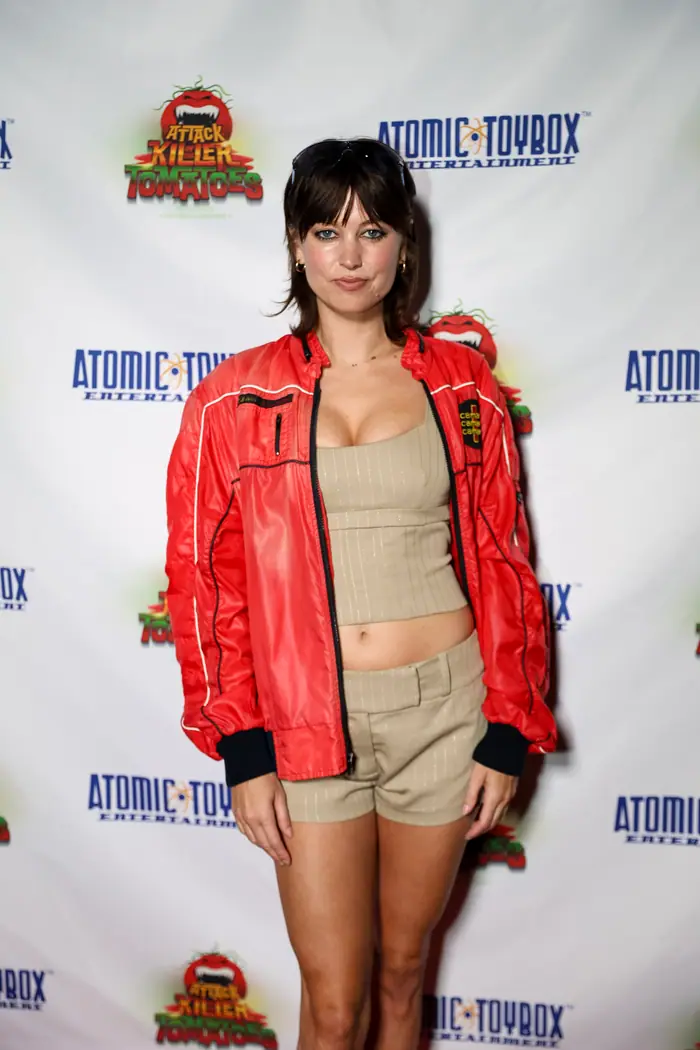 A guest in a red bomber jacket and tan shorts standing full length on the red carpet.