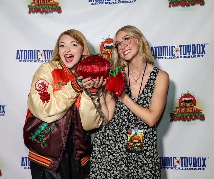 Two attendees holding up a sequinned tomato handbag, one wearing a Killer Tomatoes varsity jacket.