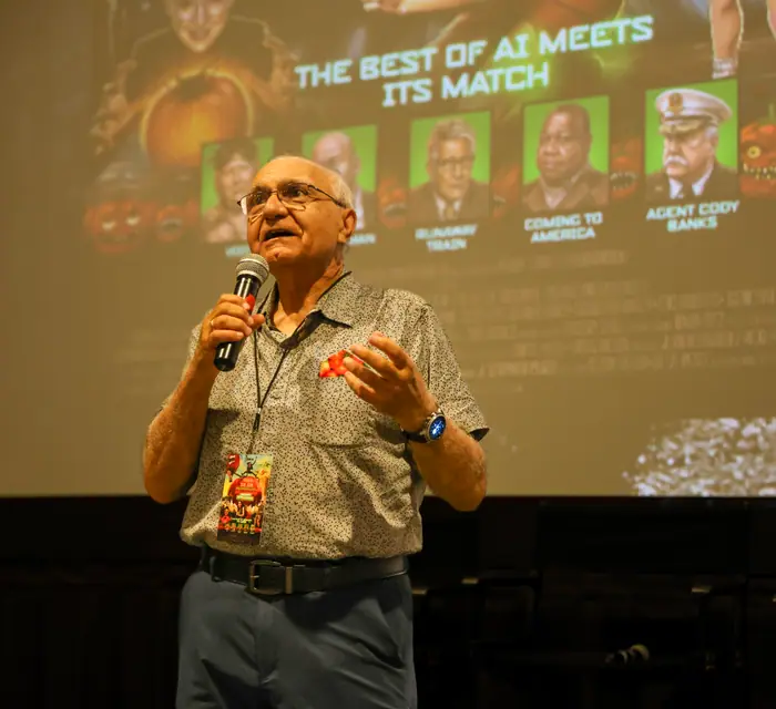 A speaker at the microphone in front of the cinema screen, mid-gesture, the film titles projected behind.