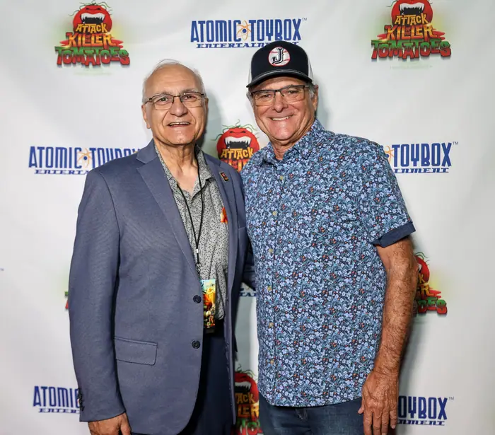 Two guests standing together on the red carpet, one in a blazer and lanyard, one in a patterned shirt and cap.